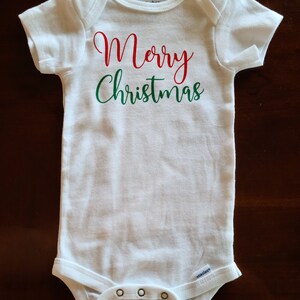 Op de afbeelding: Witte baby romper met de tekst "Merry Christmas" in rode en groene letters.