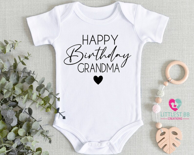 May include: White baby onesie with black text that reads "Happy Birthday Grandma" with a small black heart below the text.