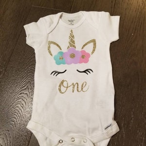 May include: White baby onesie with a gold glitter unicorn design and the word "one" in gold glitter. The unicorn has a pink, purple, and blue flower crown.