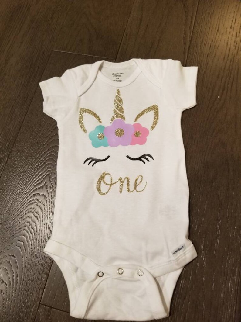 May include: White baby onesie with a gold glitter unicorn design and the word "one" in gold glitter. The unicorn has a pink, purple, and blue flower crown.