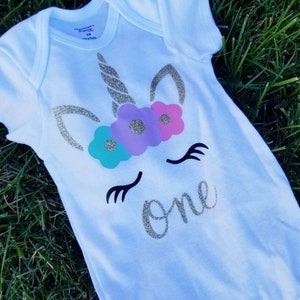 May include: White onesie with a gold glitter unicorn face and flowers. The unicorn has a gold glitter horn and closed eyes. The text "one" is written in gold glitter below the unicorn.