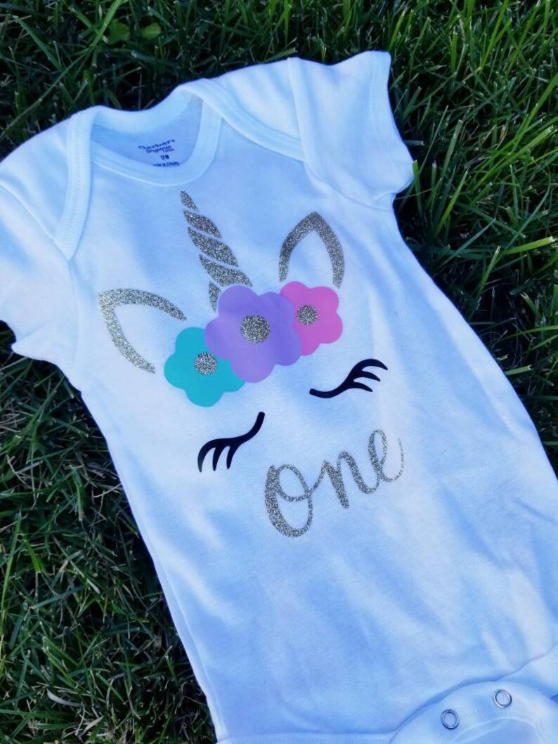 May include: White onesie with a gold glitter unicorn face and flowers. The unicorn has a gold glitter horn and closed eyes. The text "one" is written in gold glitter below the unicorn.