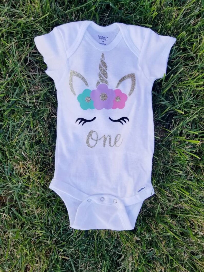 May include: White onesie with a gold glitter unicorn design and the word "one" in gold glitter. The unicorn has a pink, purple, and teal flower crown.