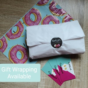 May include: A white package wrapped in tissue paper with a donut-patterned mailing bag. A sticker with the text "thank you for your business" is on the package. Three business cards with the text "Littlest Creations" are stacked on a wooden surface. A teal blue sign with the text "Gift Wrapping Available" is in the lower left corner of the image.
