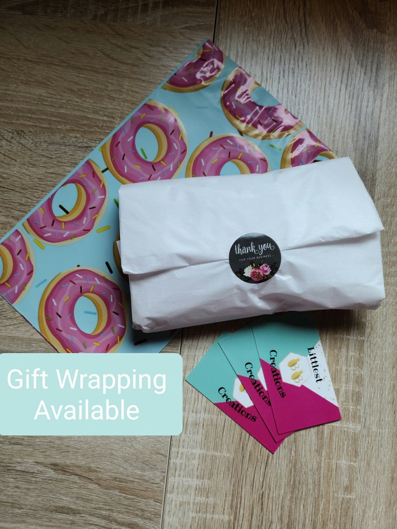 May include: A white package wrapped in tissue paper with a donut-patterned mailing bag. A sticker with the text "thank you for your business" is on the package. Three business cards with the text "Littlest Creations" are stacked on a wooden surface. A teal blue sign with the text "Gift Wrapping Available" is in the lower left corner of the image.