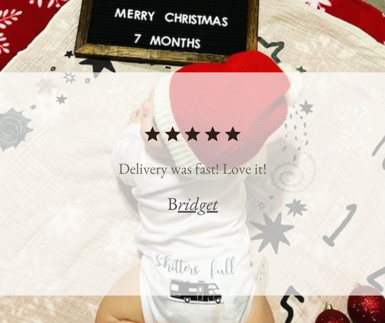 Op de afbeelding: Een witte baby romper met de tekst "Shitters full" en een zwart-witte illustratie van een camper. De romper wordt gedragen door een baby die een rode gebreide muts draagt. De baby ligt op een wit deken met een zwart letterbord waarop "Merry Christmas 7 Months" staat.