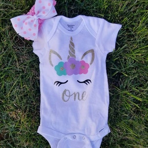 May include: White onesie with a gold glitter unicorn design and the word "one" in gold glitter. The unicorn has a pink, purple, and teal flower crown. The onesie is paired with a pink and white polka dot headband.