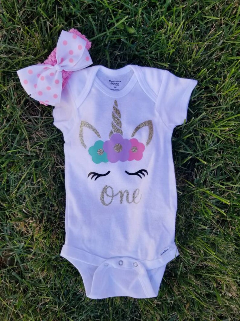 May include: White onesie with a gold glitter unicorn design and the word "one" in gold glitter. The unicorn has a pink, purple, and teal flower crown. The onesie is paired with a pink and white polka dot headband.