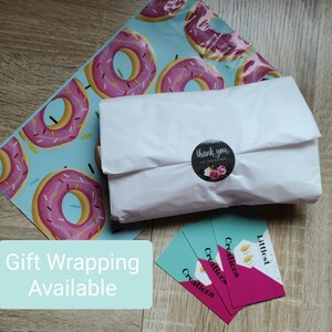 May include: A white package wrapped in tissue paper with a donut-patterned mailing bag. A sticker with the text "thank you for your business" is on the package. Three business cards with the text "Littlest Creations" are stacked on a wooden surface. A turquoise blue sign with the text "Gift Wrapping Available" is in the lower left corner of the image.