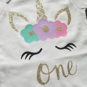 May include: White onesie with a gold glitter unicorn design. The unicorn has a gold horn, pink, purple, and teal flowers, and black eyelashes. The word "one" is written in gold glitter below the unicorn.