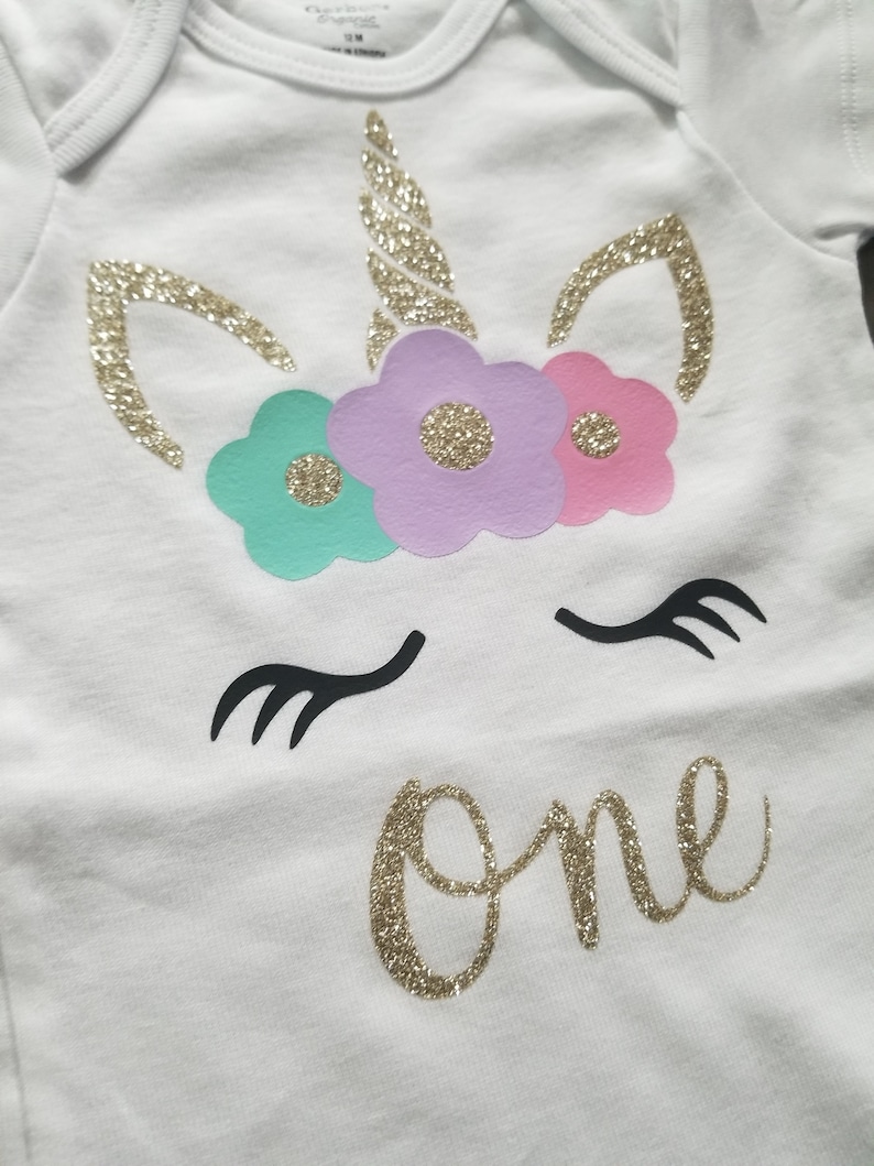 May include: White onesie with a gold glitter unicorn design. The unicorn has a gold horn, pink, purple, and teal flowers, and black eyelashes. The word "one" is written in gold glitter below the unicorn.