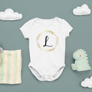May include: White baby onesie with a gold wreath and the letter L in black. The onesie is surrounded by a white cloud, a green dinosaur toy, a gray elephant toy, and a striped blanket.
