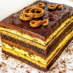 May include: A multi-layered chocolate cake with a rich chocolate frosting and a decorative swirl of caramel on top. The cake is dusted with cocoa powder and is sitting on a white surface.