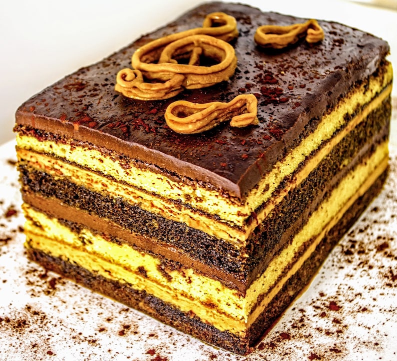 May include: A multi-layered chocolate cake with a rich chocolate frosting and a decorative swirl of caramel on top. The cake is dusted with cocoa powder and is sitting on a white surface.