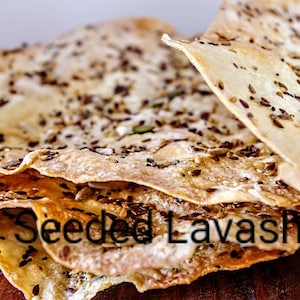 May include: A stack of thin, crispy lavash bread topped with a variety of seeds. The bread is light brown in color and has a slightly golden hue. The seeds are a mix of black, white, and brown, and they are scattered evenly across the surface of the bread. The bread is sitting on a wooden surface. "Seeded Lavash" is written in black text on the bread.