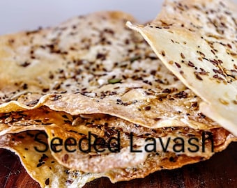 May include: A stack of thin, crispy lavash bread topped with a variety of seeds. The bread is light brown in color and has a slightly golden hue. The seeds are a mix of black, white, and brown, and they are scattered evenly across the surface of the bread. The bread is sitting on a wooden surface. "Seeded Lavash" is written in black text on the bread.
