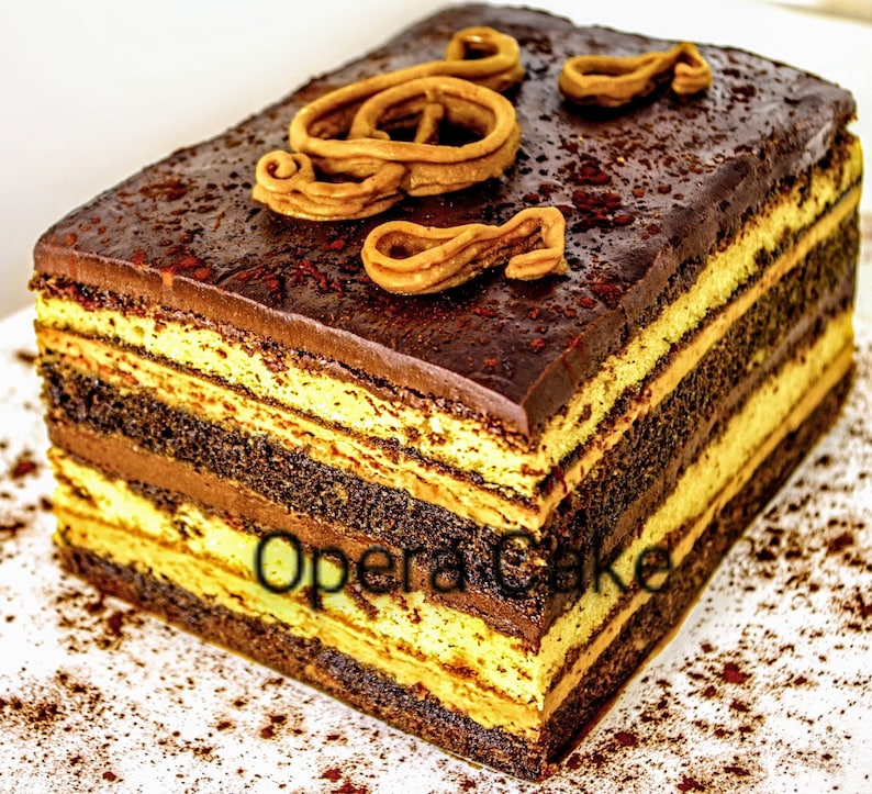 May include: A layered chocolate cake with a coffee and chocolate filling, topped with a chocolate ganache and a swirl of caramel. The cake is dusted with cocoa powder and the text "Opera Cake" is visible on the side.