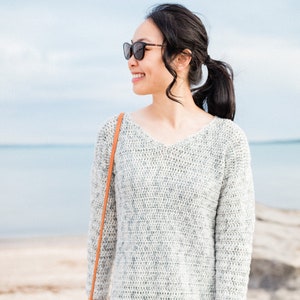 Puede incluir: Una mujer con un suéter de crochet gris y blanco con un bolso de cuero marrón con correa. Lleva pantalones negros y gafas de sol.
