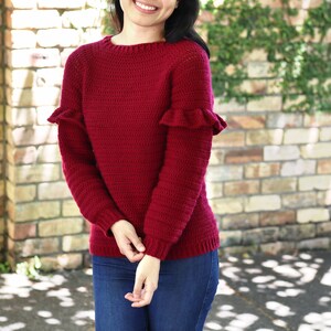 Puede incluir: Un suéter de crochet color granate con mangas con volantes. El suéter tiene un cuello redondo y mangas largas.