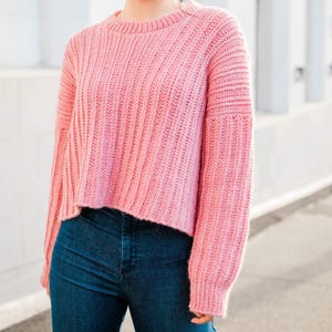 May include: A pink knitted sweater with a ribbed texture and long sleeves. The sweater is cropped and has a relaxed fit.