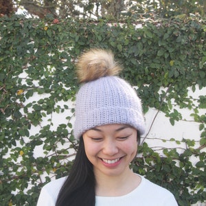 May include: A light purple knitted beanie with a brown faux fur pom pom. The beanie is being worn by a person with long black hair.