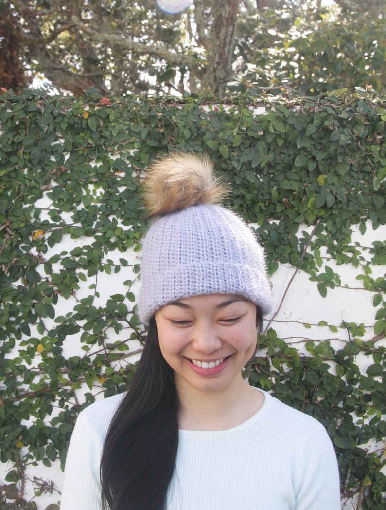 May include: A light purple knitted beanie with a brown faux fur pom pom. The beanie is being worn by a person with long black hair.