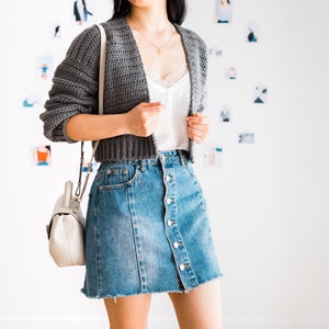 May include: A woman wearing a gray cropped cardigan over a white tank top and a denim skirt with buttons down the front. She is carrying a white handbag with a strap.
