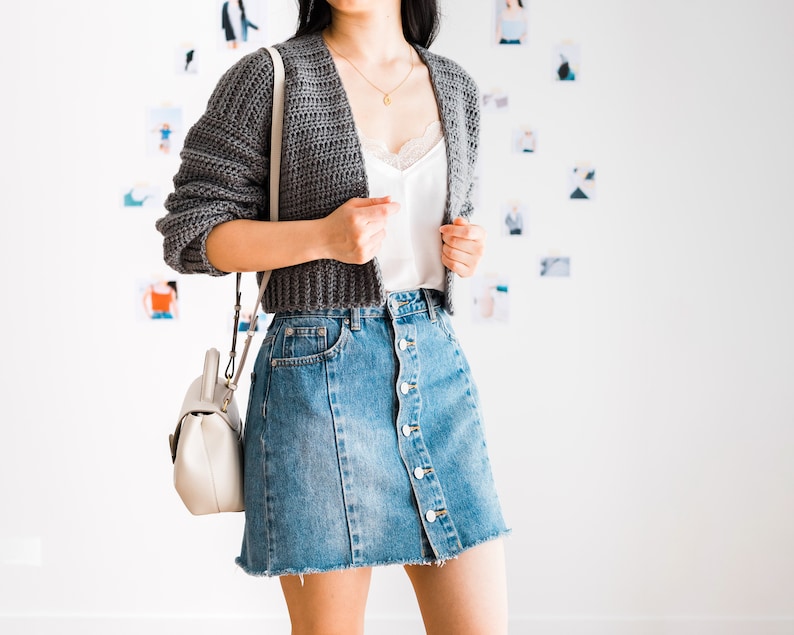 May include: A woman wearing a gray cropped cardigan over a white tank top and a denim skirt with buttons down the front. She is carrying a white handbag with a strap.