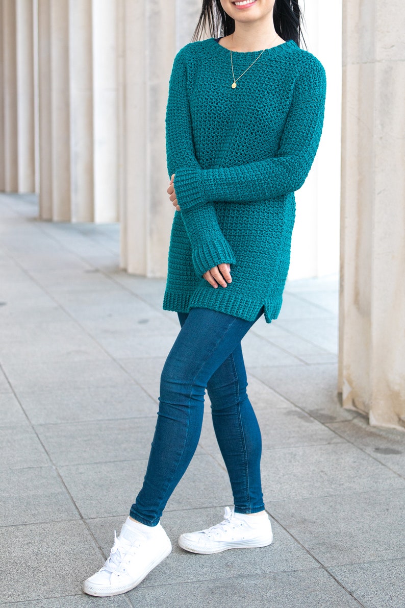 May include: A teal blue crocheted sweater with long sleeves and a relaxed fit. The sweater is worn with blue jeans and white sneakers.