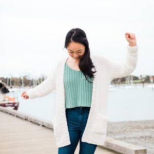 May include: A woman wearing a white crocheted cardigan over a light green crocheted top and blue jeans. She is standing on a wooden jetty with water in the background.