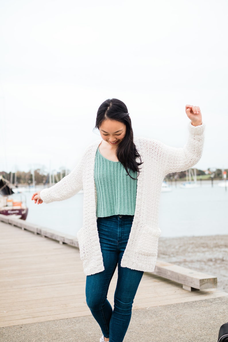 May include: A woman wearing a white crocheted cardigan over a light green crocheted top and blue jeans. She is standing on a wooden jetty with water in the background.
