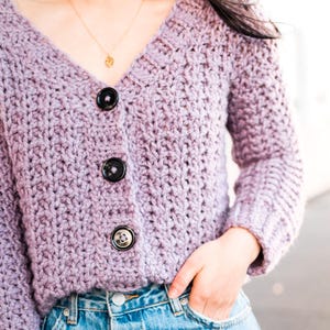 May include: A purple knitted cardigan with a v-neck and three black buttons. The cardigan is worn over a pair of blue denim jeans.