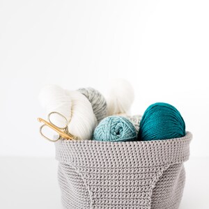 May include: A gray crocheted basket filled with balls of yarn in white, gray, teal, and turquoise. A pair of gold crochet hooks are also in the basket.
