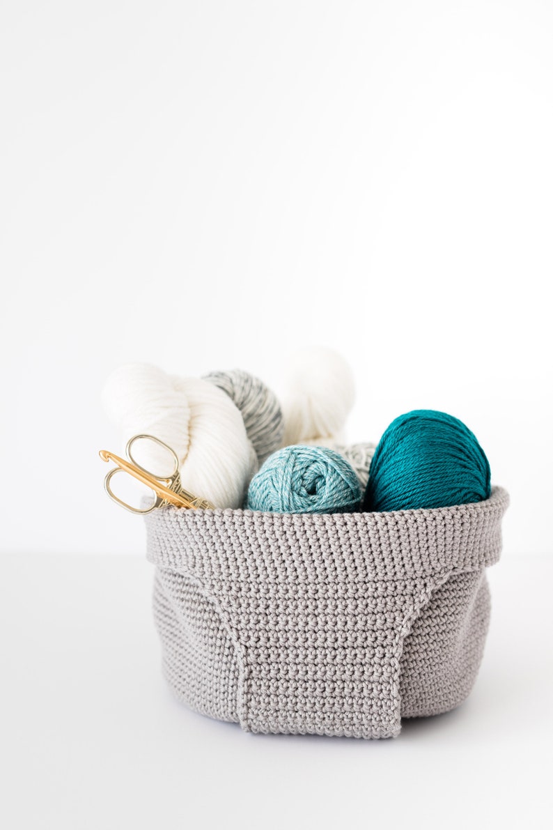 May include: A gray crocheted basket filled with balls of yarn in white, gray, teal, and turquoise. A pair of gold crochet hooks are also in the basket.