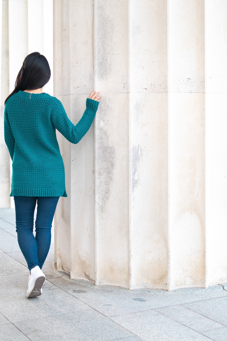 May include: A woman wearing a teal blue sweater and blue jeans stands in front of a white pillar.