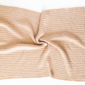 May include: A beige knitted throw blanket with a textured weave. The blanket is folded in half and has a soft, cosy appearance.