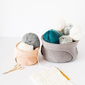 May include: Two crocheted baskets filled with balls of yarn in various colors, including teal, gray, and white. A crochet hook and scissors are also pictured.