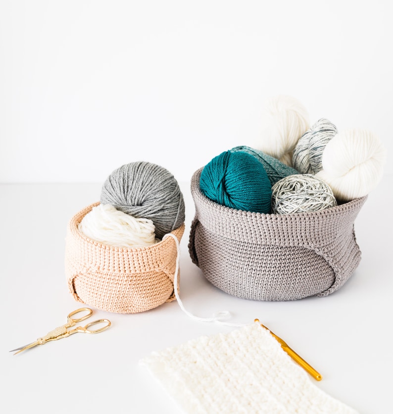 May include: Two crocheted baskets filled with balls of yarn in various colors, including teal, gray, and white. A crochet hook and scissors are also pictured.