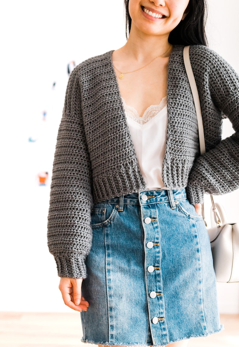 May include: A gray cropped cardigan with long sleeves and a V-neckline. The cardigan is worn over a white tank top with lace trim and a denim skirt with buttons down the front.
