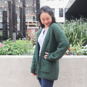 May include: A dark green crocheted cardigan with long sleeves and pockets. The cardigan is worn by a person with dark hair and is standing in front of a concrete wall.
