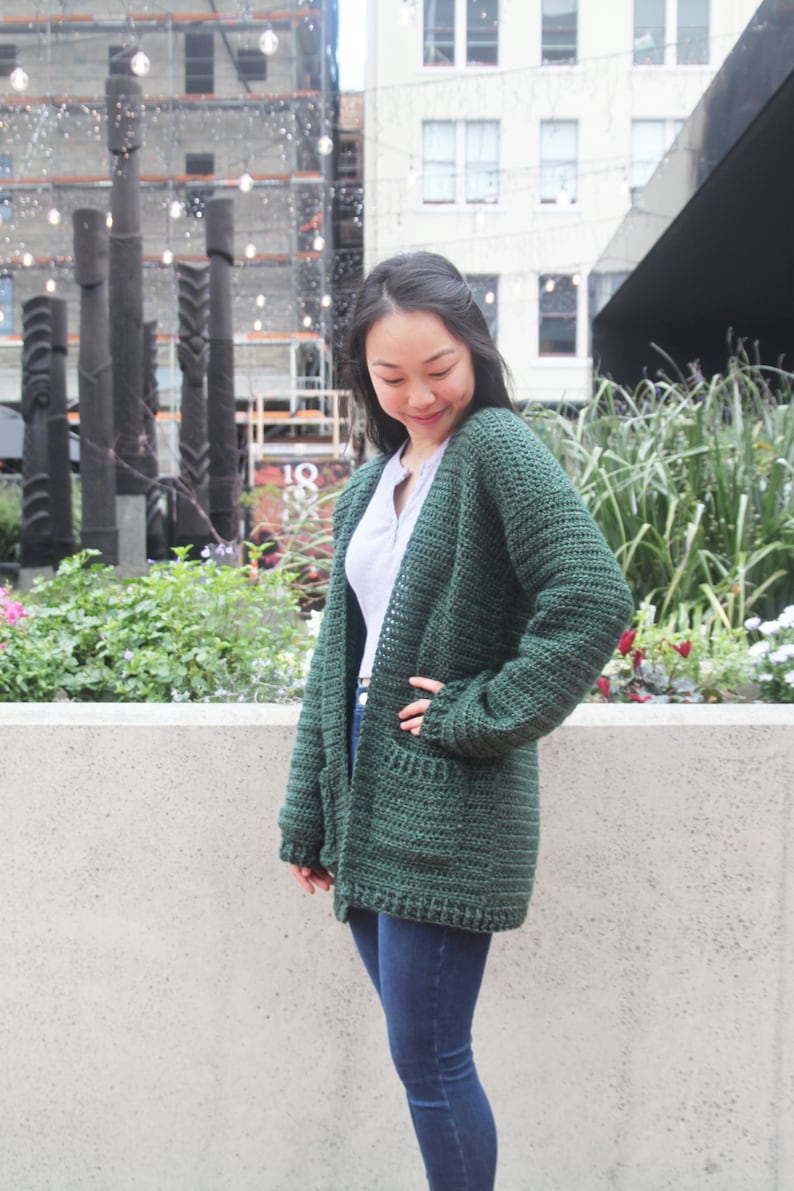 May include: A dark green crocheted cardigan with long sleeves and pockets. The cardigan is worn by a person with dark hair and is standing in front of a concrete wall.