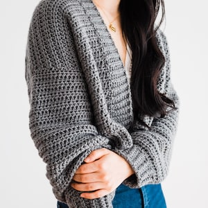 May include: A gray crocheted cardigan with a v-neck and long sleeves. The cardigan is worn by a person with long black hair.