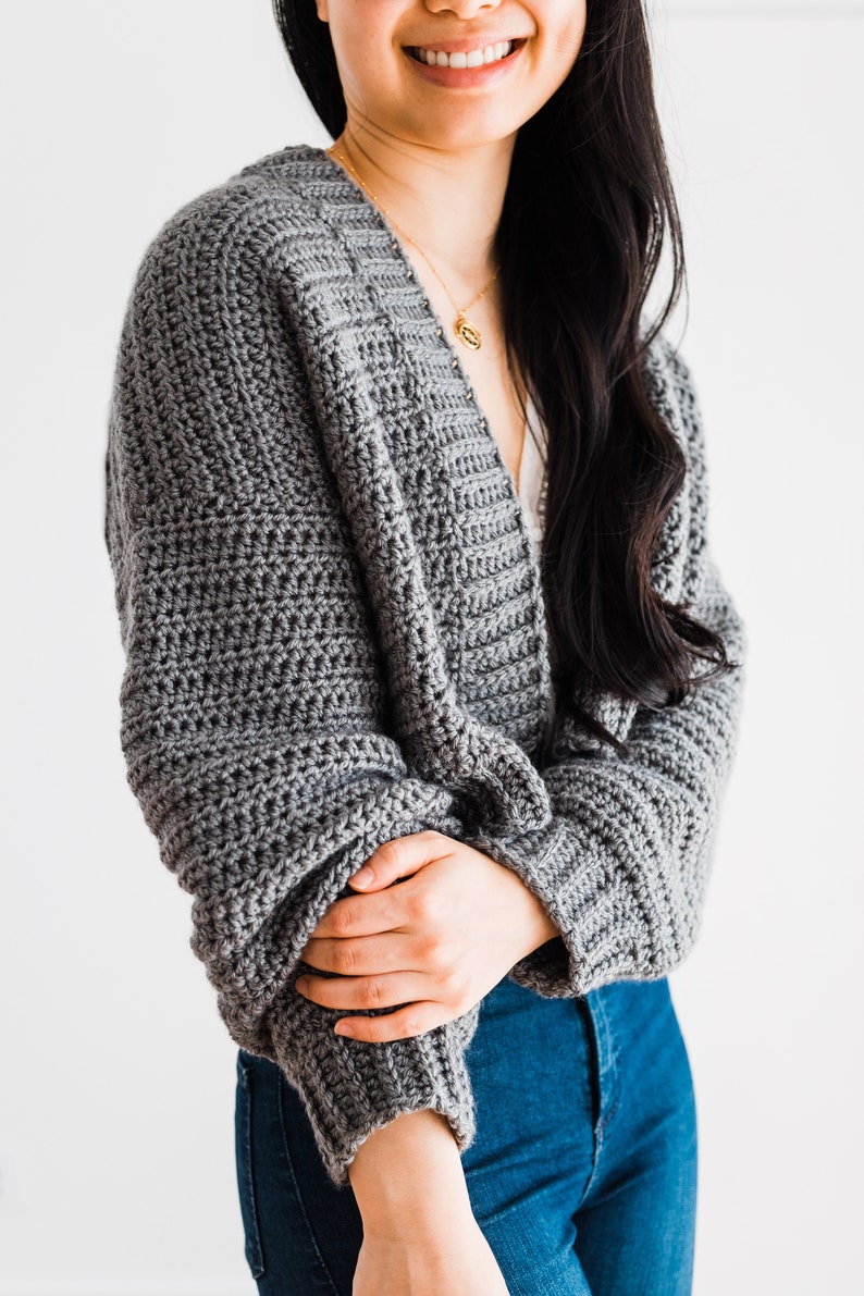 May include: A gray crocheted cardigan with a v-neck and long sleeves. The cardigan is worn by a person with long black hair.