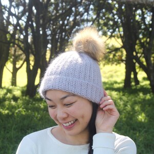 May include: A woman wearing a light purple knitted beanie with a brown faux fur pom pom. The beanie has a ribbed texture. She is smiling and looking down.