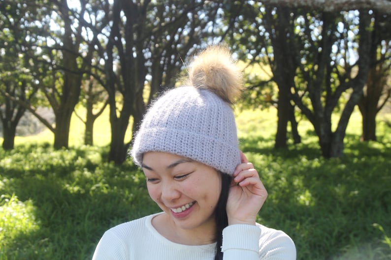 May include: A woman wearing a light purple knitted beanie with a brown faux fur pom pom. The beanie has a ribbed texture. She is smiling and looking down.