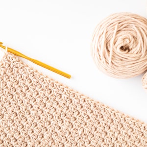 May include: A close-up of a beige crochet project in progress with a gold crochet hook and two balls of beige yarn.
