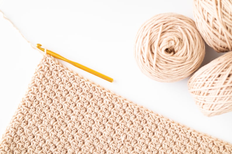 May include: A close-up of a beige crochet project in progress with a gold crochet hook and two balls of beige yarn.