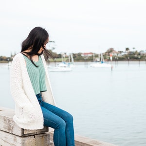 May include: A woman wearing a white cardigan, a light green tank top, and blue jeans sits on a wooden pier overlooking a body of water with boats in the distance.
