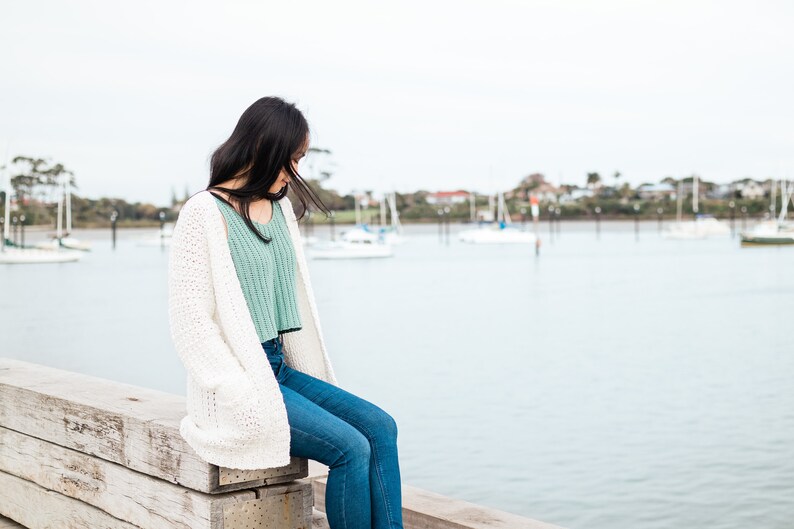 May include: A woman wearing a white cardigan, a light green tank top, and blue jeans sits on a wooden pier overlooking a body of water with boats in the distance.
