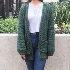 May include: A dark green crocheted cardigan with long sleeves and two pockets. The cardigan is open in the front and has a relaxed fit.